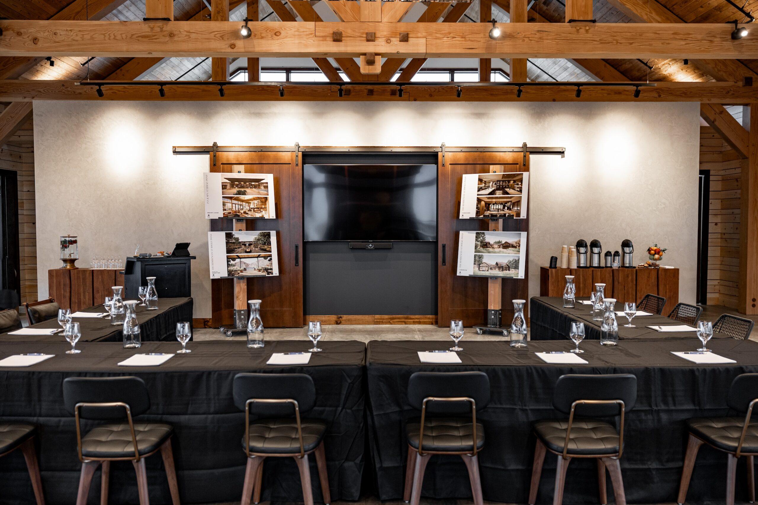 A conference room with wooden beams and modern decor, set up with black tablecloths, glassware, notepads, and chairs facing a large screen. Posters with architectural designs are displayed on either side.